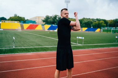 Genç, modaya uygun siyah eşofmanlı sporcu vücudunun üst kısımları için egzersiz yapıyor. Sağlıklı yaşam tarzını korumak için gülümsüyor, yakışıklı erkek koşucu kollarını çalıştırıyor ve vücut çalışmalarının tadını çıkarıyor.