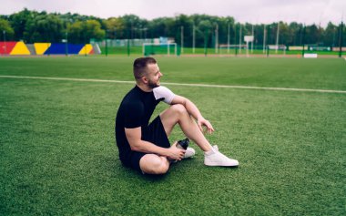 Spor kıyafetleri içinde, yeşil çimlerde elinde su şişesiyle oturan neşeli, genç bir erkek sporcunun yan görüntüsü.