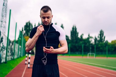 Aktif kıyafetli, kulaklık takmış, elinde akıllı telefonuyla stadyumdaki koşu pistinde duran yakışıklı bir erkek sporcu.