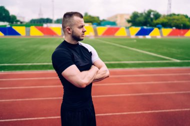 Ciddi bir beyaz erkek antrenör sabah antrenmanında elleri çapraz şekilde stadyumda duruyor. 20 'li yaşlarda yakışıklı, dalgın sporcu gözlerini kaçırarak antrenmandan memnun.