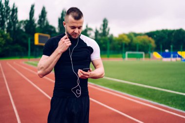 Modern kulaklıklarda stadyumdaki akıllı telefon çalma listesinden müzik seçen beyaz erkek koşucu, cep telefonundaki uygulamadan memnun olan pozitif meloman sporcusu