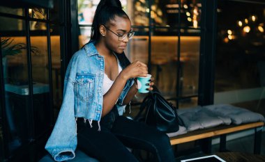 Konsantre modaya uygun giyinmiş gözlüklü siyah kadın cam duvarın yanındaki göz kamaştırıcı bankta oturuyor ve kahvesinde şeker karıştırıyor.
