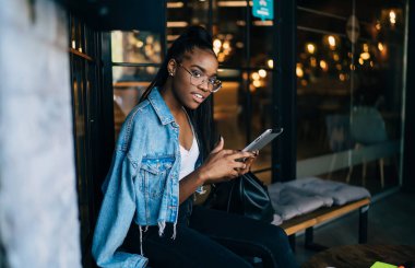 Modayı takip eden esmer tenli güzel bir kadının yarım boy portresi kafenin içinde boş zamanlarında modern cihazlar kullanıyor, pozitif Afro-Amerikan kadını dijital tablet tutuyor ve multimedya içeriğini paylaşıyor.