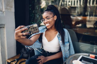 Moda giysiler içinde neşeli koyu tenli bir kadın kafeterya terasında oturmuş fotoğraf çekiyor ve blog için medya içeriği hazırlıyor, Afro-Amerikan kadın 20 'li yaşlarda cep telefonu kullanarak selfie çekiyor.