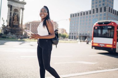 Elinde dijital tabletle gülümseyen ve müzik dinlemekten zevk alan genç, esmer bir kadın şehir ortamlarında yürüyen neşeli Afrikalı Amerikalı hippi kız dokunmatik pedi ve kulaklıkla kasabada yürüyor.