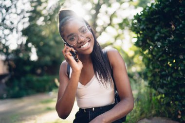 Parktaki yeşil çalıların yanında otururken gözlüklü, gülümseyen ve cep telefonuyla konuşan iyimser bir bayan.
