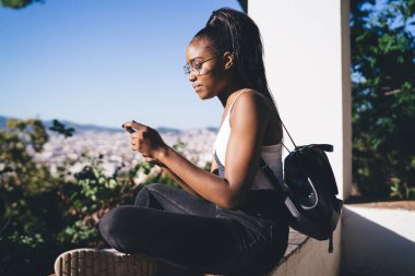 Güneşli bir günde ağaçların arasından şehrin manzarasının keyfini çıkarırken ekrandaki kadın akıllı telefona odaklanmış siyahın yan görüntüsü