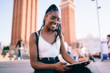 Moda tişörtlü, gözlüklü, kameraya gülümseyen ve şehir meydanında otururken tablete gülümseyen neşeli Afro-Amerikan kadının alçak açısı.