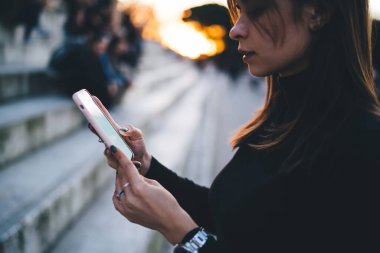 Günbatımına karşı sokakta dikilirken günlük kıyafetleriyle akıllı telefonları karıştıran genç ve ciddi bir kadının yan görüntüsü