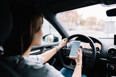 Şehir caddesinde araba sürerken sıradan giysiler içinde oturan ve akıllı telefonlara bakan tanınmayan genç bir kadının arka görüntüsü.