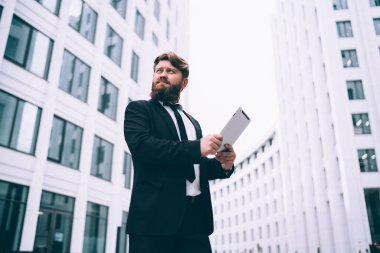 Ciddi, tombul, tıraşsız, akıllı takım elbiseli, elinde tabletle modern şehir binalarının arasında duran ve gözlerini kaçıran erkek girişimcinin düşük açısı.