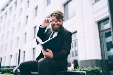 Resmi takım elbiseli mutlu sakallı iş adamının elinde belgelerle ahşap bankta oturup kameraya bakarken gülümsemesinden.