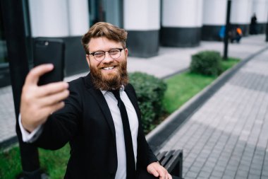 Resmi takım elbiseli, sakallı ve gözlüklü neşeli genç girişimci ofis binasının avlusunda otururken selfie çekiyor.