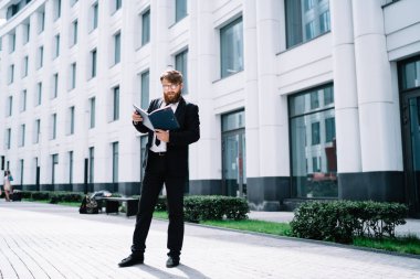 Resmi takım elbiseli ciddi bir işadamının tam vücudu ofis binasının yakınında dikilirken belgeleri okuyor.