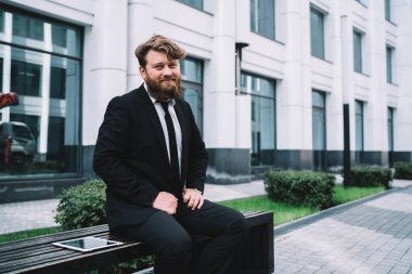 İyi giyimli, sakallı bir ofis çalışanı meydanda oturuyor ve dinlenirken gülümseyerek kameraya bakıyor.