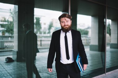 Modern cam duvarın yanında duran ve kameraya bakarken klasörleri elinde tutan modaya uygun takım elbiseli başarılı bir iş adamı.