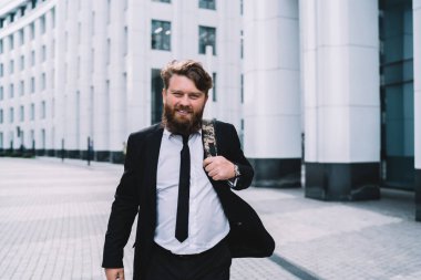 Resmi takım elbiseli, sakallı mutlu genç adam işten eve dönerken parlak dişli gülüşlü kameraya bakıyor.