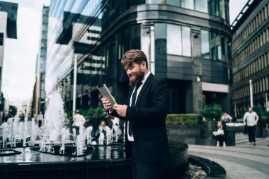 Resmi takım elbiseli ve kravatlı içerik sakallı işadamının sokak çeşmesinin yanında durup şehirdeki tableti karıştırması...