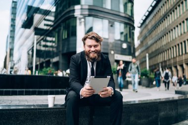 Resmi kıyafetli ve kravatlı mutlu sakallı erkek yönetici taş sınırında oturup kameraya bakarken gülümsüyor ve tabletle takılıyor.