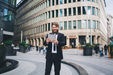 Resmi kıyafetli, gür sakallı ve neşeli gülümsemeli genç adam modern şehir parkında kahve içerken heyecanla tablet izliyor.