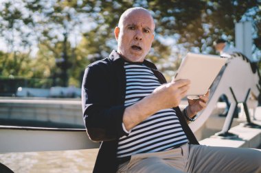 Gri sakallı, sıradan giyimli, elinde tabletle parkta göletin kenarında otururken kameraya bakıp konuşan yaşlı adama konsantre ol.