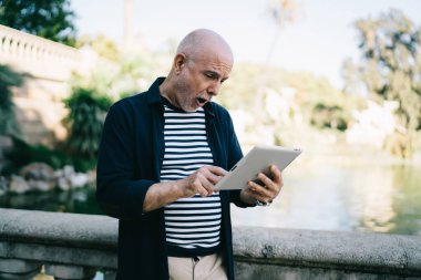 Şehir parkında tırabzanın yanında dikilen ve tablet üzerinde şaşırmış bir şekilde duran mutlu kıdemli adam.