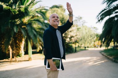 50 yaşında, elinde tabletle neşeli, yakışıklı bir erkek. Güneşli bir günde şehir parkında jest yapıyor ve gülümsüyor.