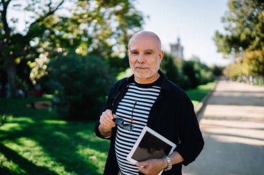 Ciddi, orta yaşlı, şık giyinmiş, güneş gözlüklü, tabletli, dost canlısı bir erkek. Park sokağındaki kameraya bakıyor.