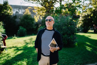 Şık giyinmiş, güneş gözlüklü kendinden emin bir erkek güneşli bir günde tabletle şehir parkında vakit geçirip gözlerini kaçırıyor.