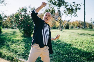 Düşük açılı modern gri saçlı erkek kulaklıkla müzik dinliyor ve kolları havada yürürken ve dans ederken şarkı söylüyor.