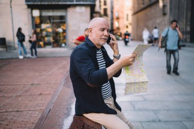 Şehir merkezinde sınırda otururken cep telefonuyla konuşan ve haritada yerini arayan yaşlı bir erkek görüntüsü