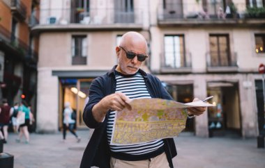From below of focused old male traveler in casual clothes and sunglasses looking through map while standing in city street clipart