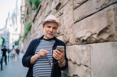 Yetişkin bir adamın caddede durup, cep telefonuyla internette sörf yaparken eski binaların tuğla duvarına yaslanmasının düşük açısı.