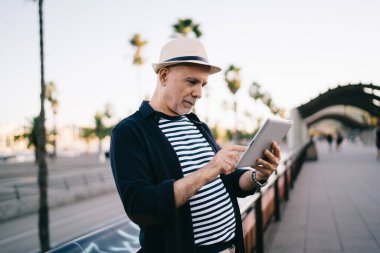 Şehirde tablet kullanarak dolaşırken parktaki tırabzanın yanında günlük kıyafetli iyimser yaşlı bir adamın yan görüntüsü.