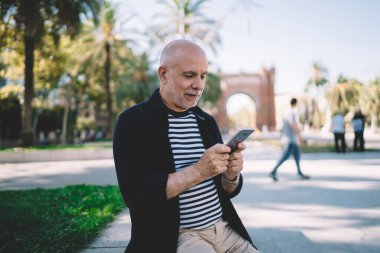 Gündelik kıyafetlerle gezen mutlu turist şehir parkında dinlenirken taştan korkuluklara oturuyor ve cep telefonuyla mesajlaşıyor.