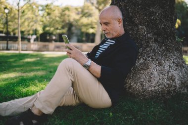 Sıradan kıyafetlerle gezen, cep telefonuyla internette sörf yapan ve parkta dinlenirken ağaca yaslanan son sınıf öğrencisinin yan görüntüsü.