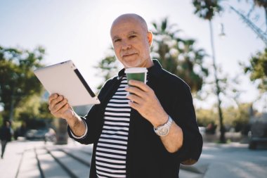 Elinde tabletle şehir parkında dururken kol saati takmış, kameraya bakıp kahvesini yudumlayan mutlu son sınıf öğrencisi.