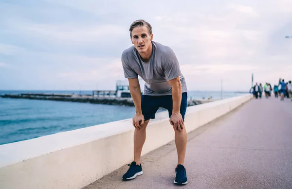 Strong young male jogger in activewear resting after cardio training legs and reaching fitness goals outdoors, sportsman having morning workout near water breathing on quay and looking at camera