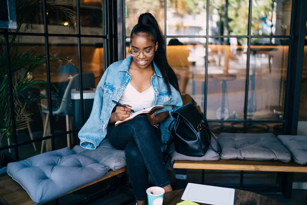 穿着休闲装的积极的黑皮肤女人坐在咖啡馆的内部享受业余时间 微笑着写东西的女性博主 并计划满足于空闲时间 — 图库照片