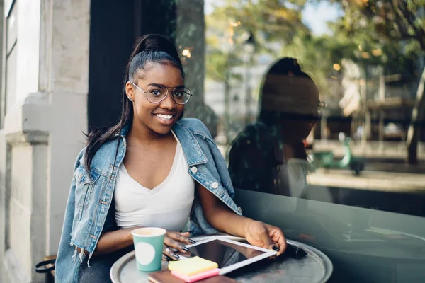 快乐的非洲裔美国女人坐在桌旁 一边用平板电脑 一边拿着黏糊糊的字条和纸杯咖啡 一边对着相机微笑 — 图库照片