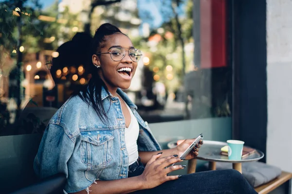 穿着牛仔夹克的快乐的非洲裔美国女士坐在现代街边咖啡店的玻璃墙边 手持智能手机和一次性咖啡 — 图库照片