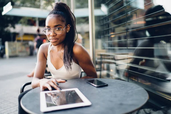 Akıllı telefon ve tabletle tatmin olmuş genç Afro-Amerikan bayan kameraya bakıyor ve düşünüyor sokak kafesinde dinlenirken ve tablet üzerinde sohbet ederken