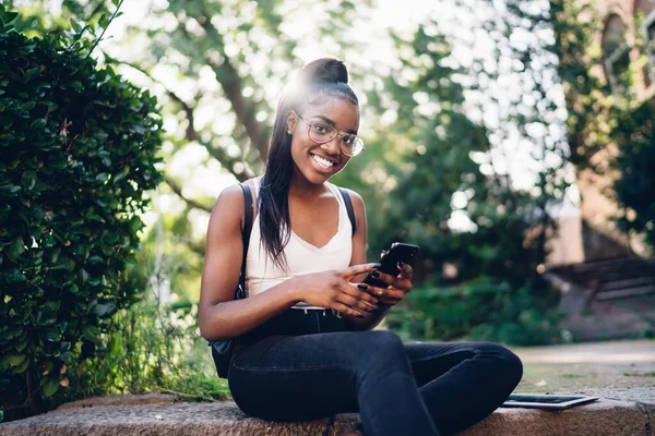 Pozemní Úroveň Usmívající Mladá Afroameričanka Ležérním Oblečení Brýlích Sedí Obrubníku — Stock fotografie