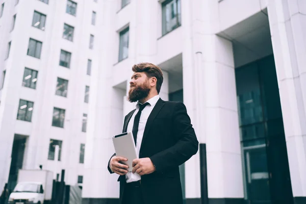 深刻なふっくらしたUnshaven男起業家の低い角度でオフィススーツでタブレットで手に立って近代的な都市の建物のファサードと離れて見て — ストック写真