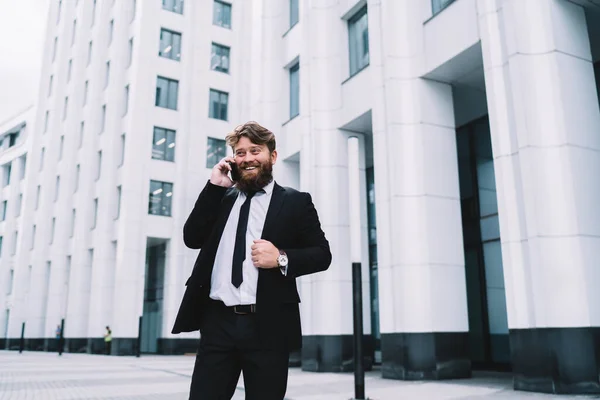 身穿雅致正装的快乐男性商人站在摩天大楼附近的大街上 一边微笑着一边用智能手机交谈 一边望着远方 — 图库照片