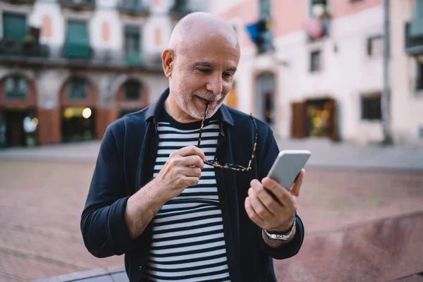 在街上站着 用手机在社交媒体上发短信时 用太阳镜对着嘴唇微笑的秃头老人 — 图库照片