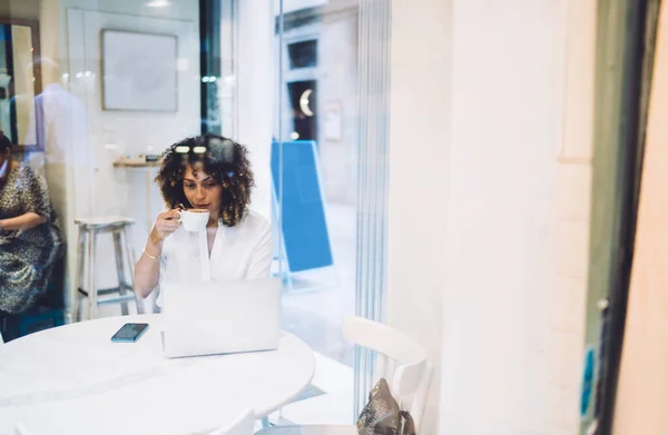 透过玻璃看身穿休闲装的年轻女企业家坐在笔记本电脑旁 喝着新鲜热咖啡 — 图库照片