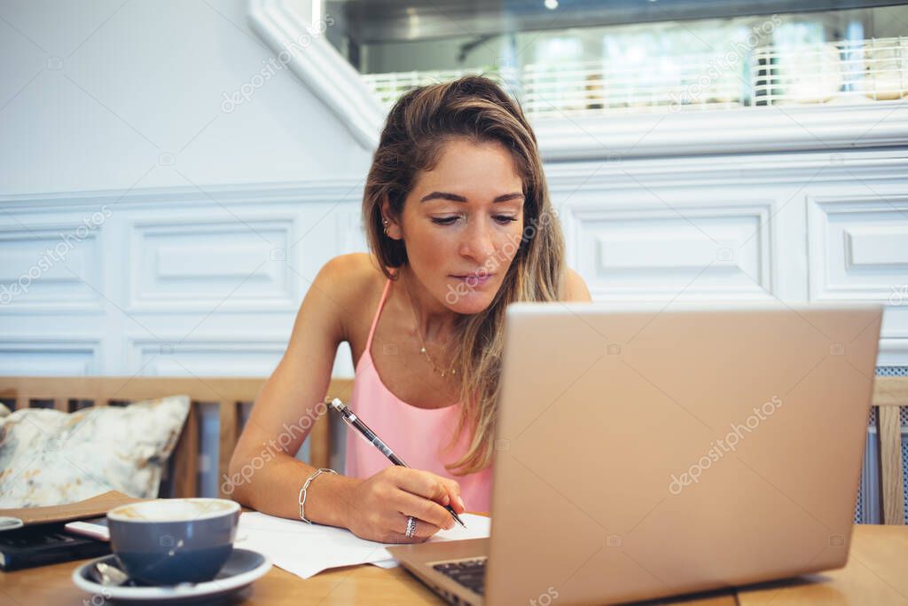 Focused female freelancer in casual clothes browsing netbook and taking notes while sitting at table and working remotely in cafe