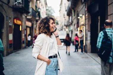Modern şehrin caddesinde kaldırımda dikilen ve akıllı telefondan kafasını çevirerek konuşan modaya uygun giyinmiş genç etnik bayanın yan görüntüsü