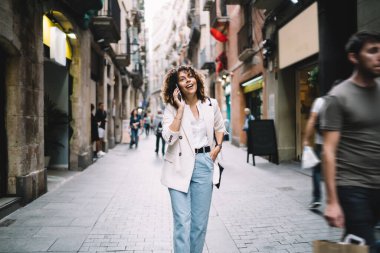 Olumlu etnik bayan gündelik giysiler içinde dar bir sokakta yürüyor ve akıllı telefondan kafasını çeviriyor.